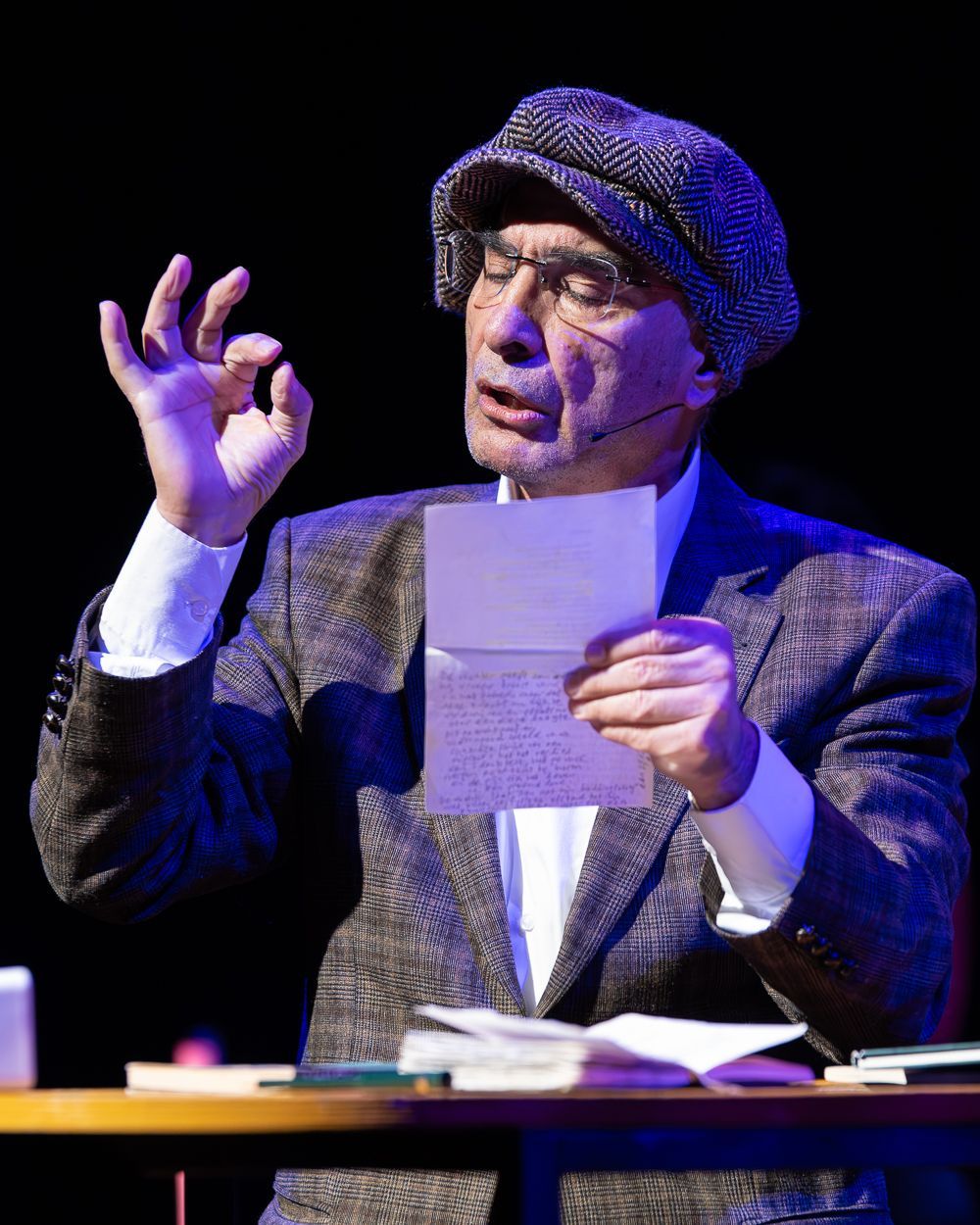 Man met papier in de hand, gebarend. Draagt een tweedjasje, pet en bril. Donkere achtergrond, bureau vol spullen.