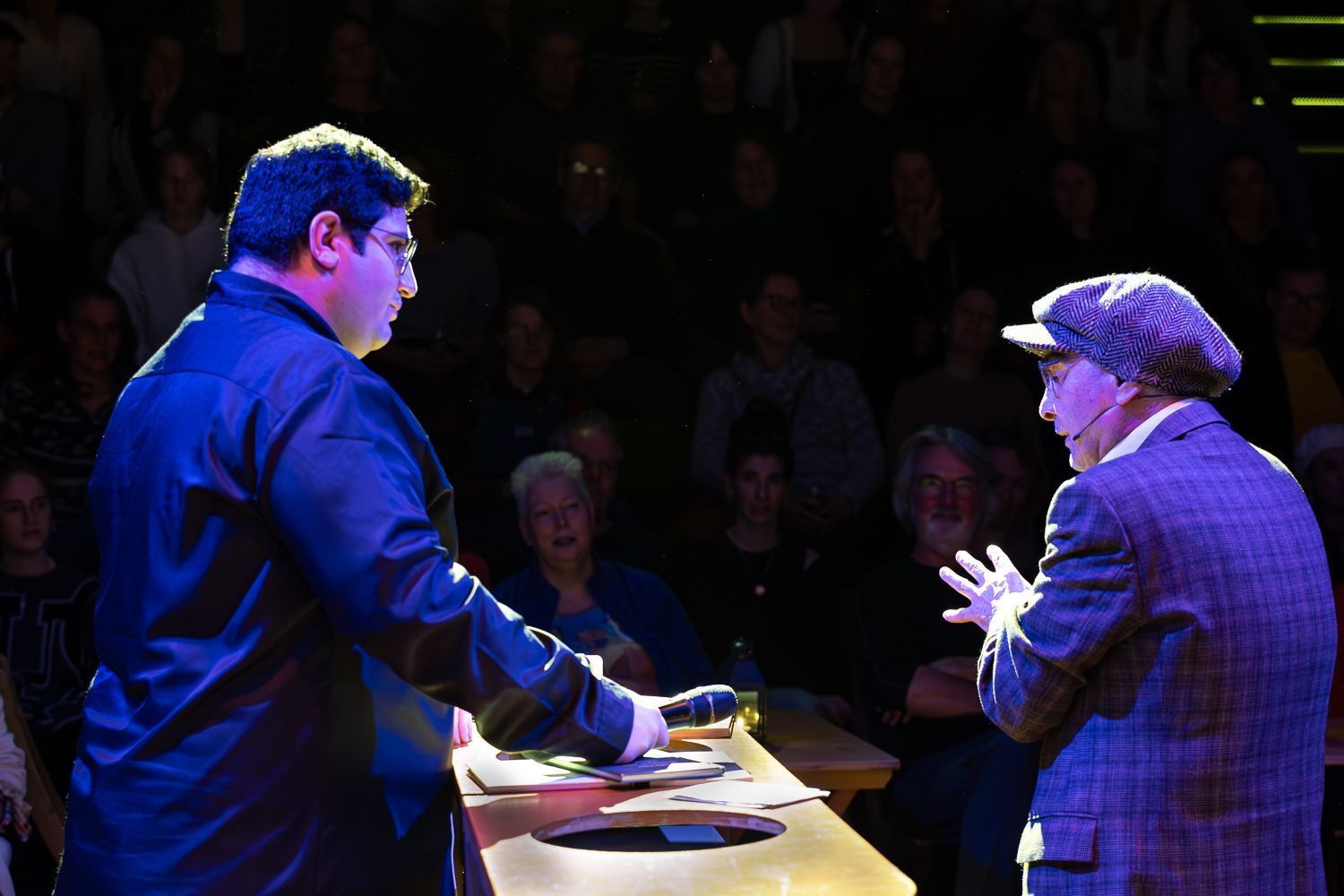 Twee mannen op het podium, één met een microfoon, tegenover elkaar. Het publiek zit op de achtergrond.