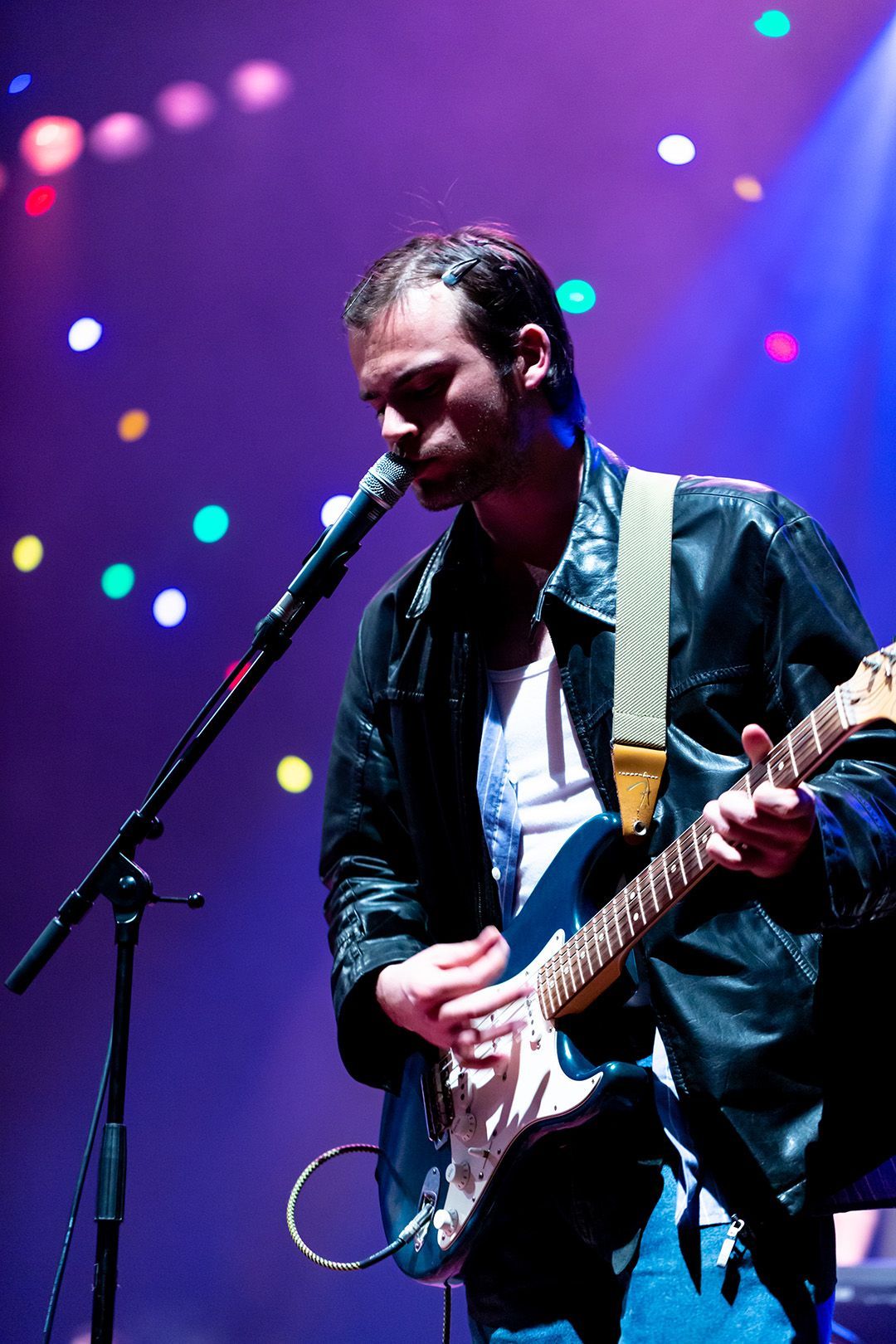 Man speelt gitaar en zingt in een microfoon op het podium, onder blauwe en kleurrijke podiumverlichting.