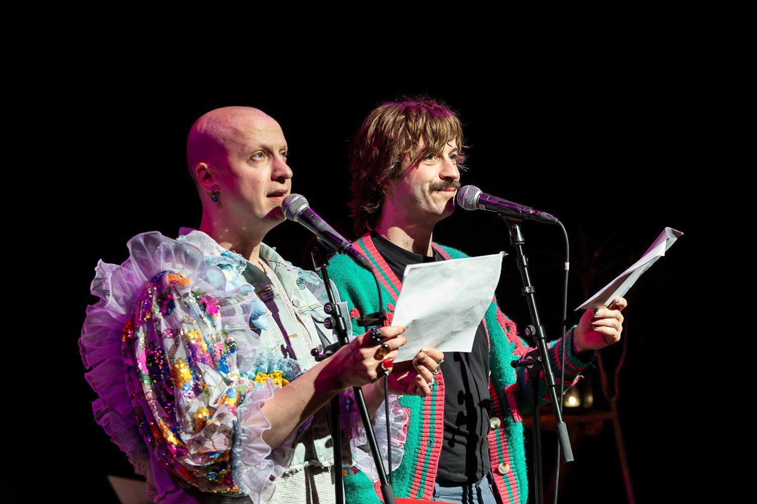 Poezieboys op het podium met tekst, de een met een kleurrijk jasje en de ander met een kaal hoofd en een topje met kant.