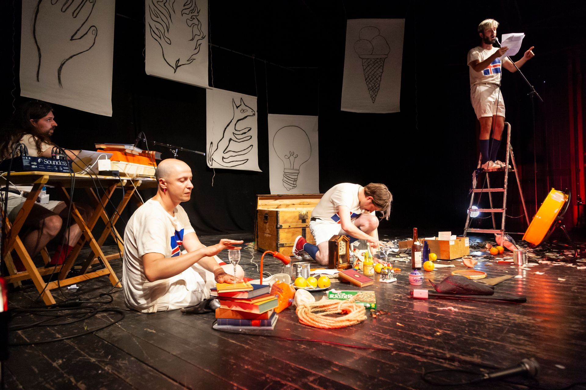 Optreden op het podium: artiesten die dingen bouwen en voorlezen, met rekwisieten, hangende kunst en lichten.