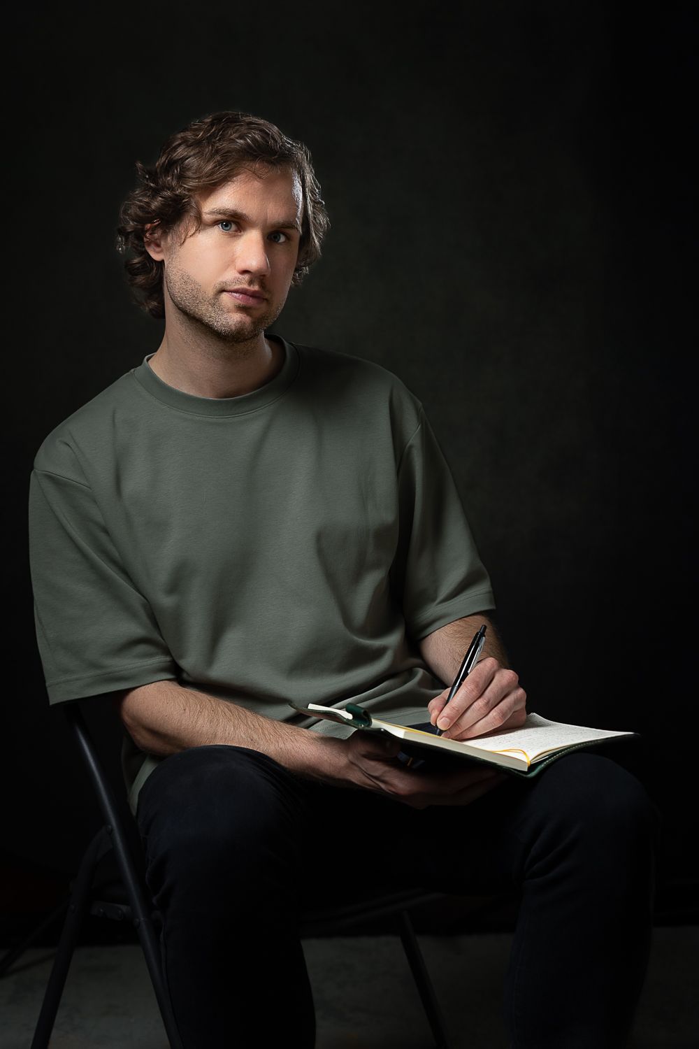 Man met krullend haar, schrijvend in een notitieblok, zittend op een zwarte stoel tegen een donkere achtergrond.