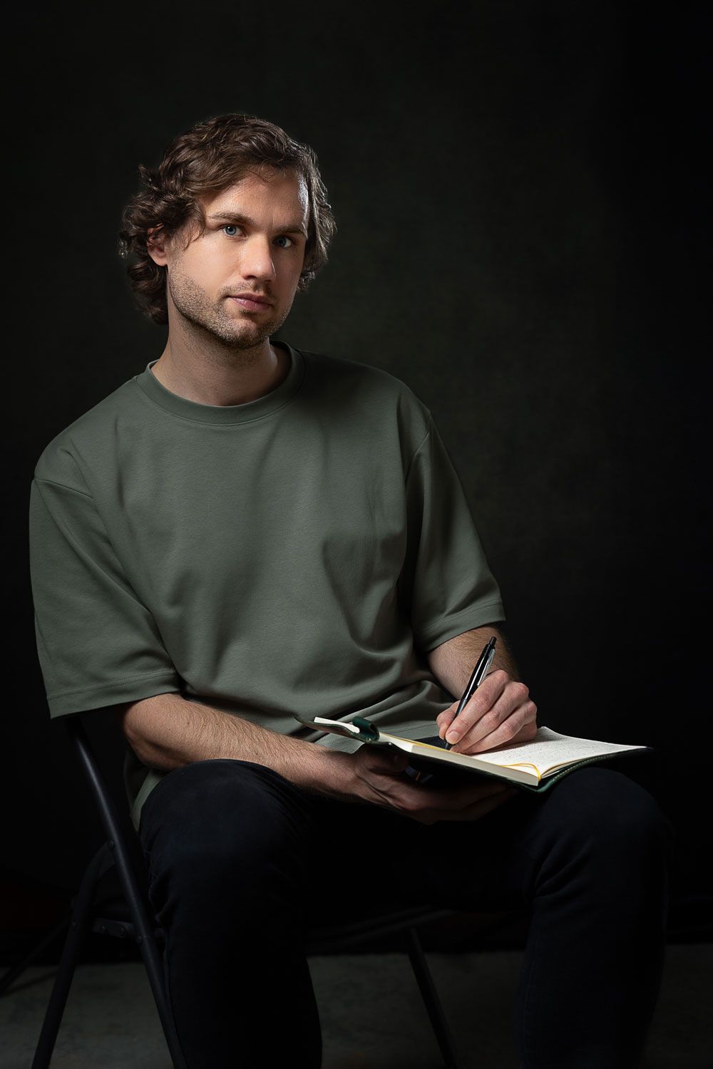 Zittende man, schrijvend in notitieboekje. Draagt ​​een groen shirt, een donkere broek. Zwarte achtergrond.