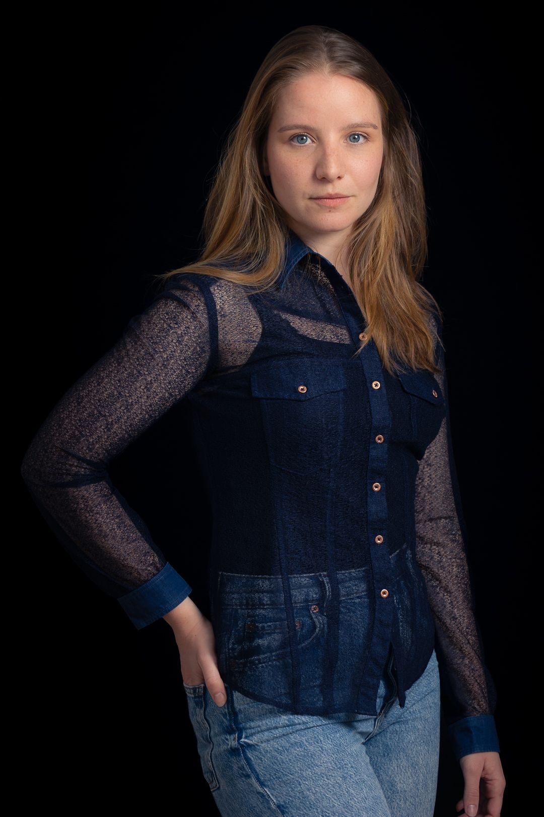 Vrouw in een blauw kanten shirt, hand in haar zak, kijkend naar de camera. Zwarte achtergrond.