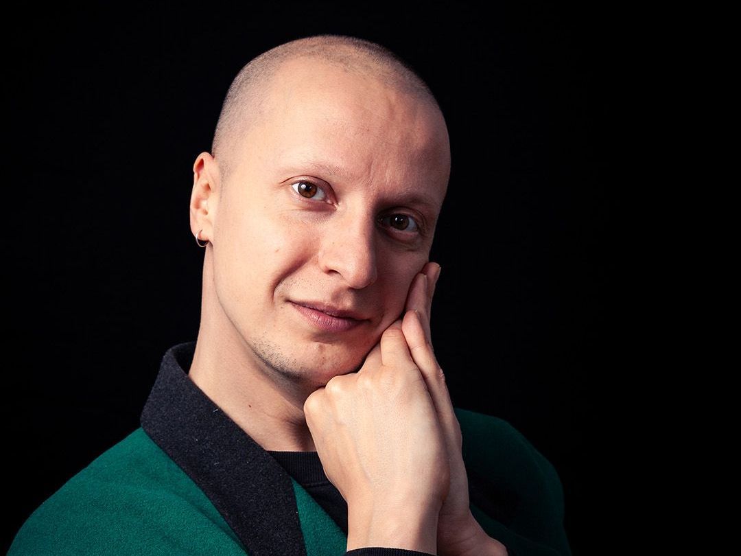 Kaal persoon in een groen shirt met handen op het gezicht, lachend tegen een zwarte achtergrond.