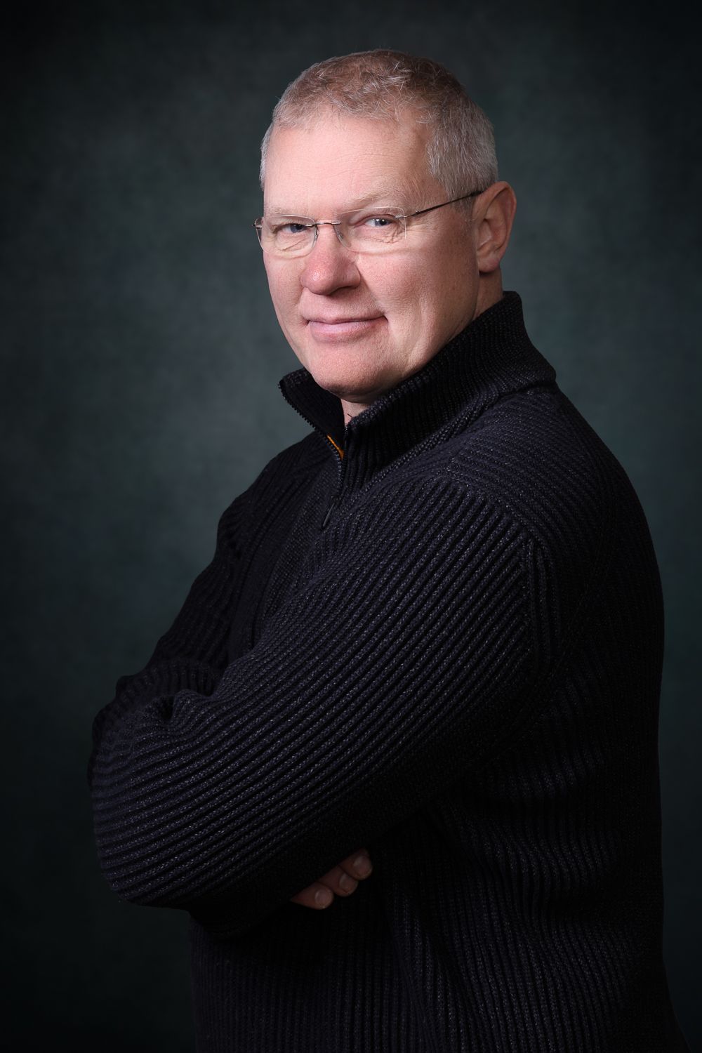 Man met bril, armen over elkaar, gekleed in een zwarte trui, staand voor een donkergroene achtergrond.