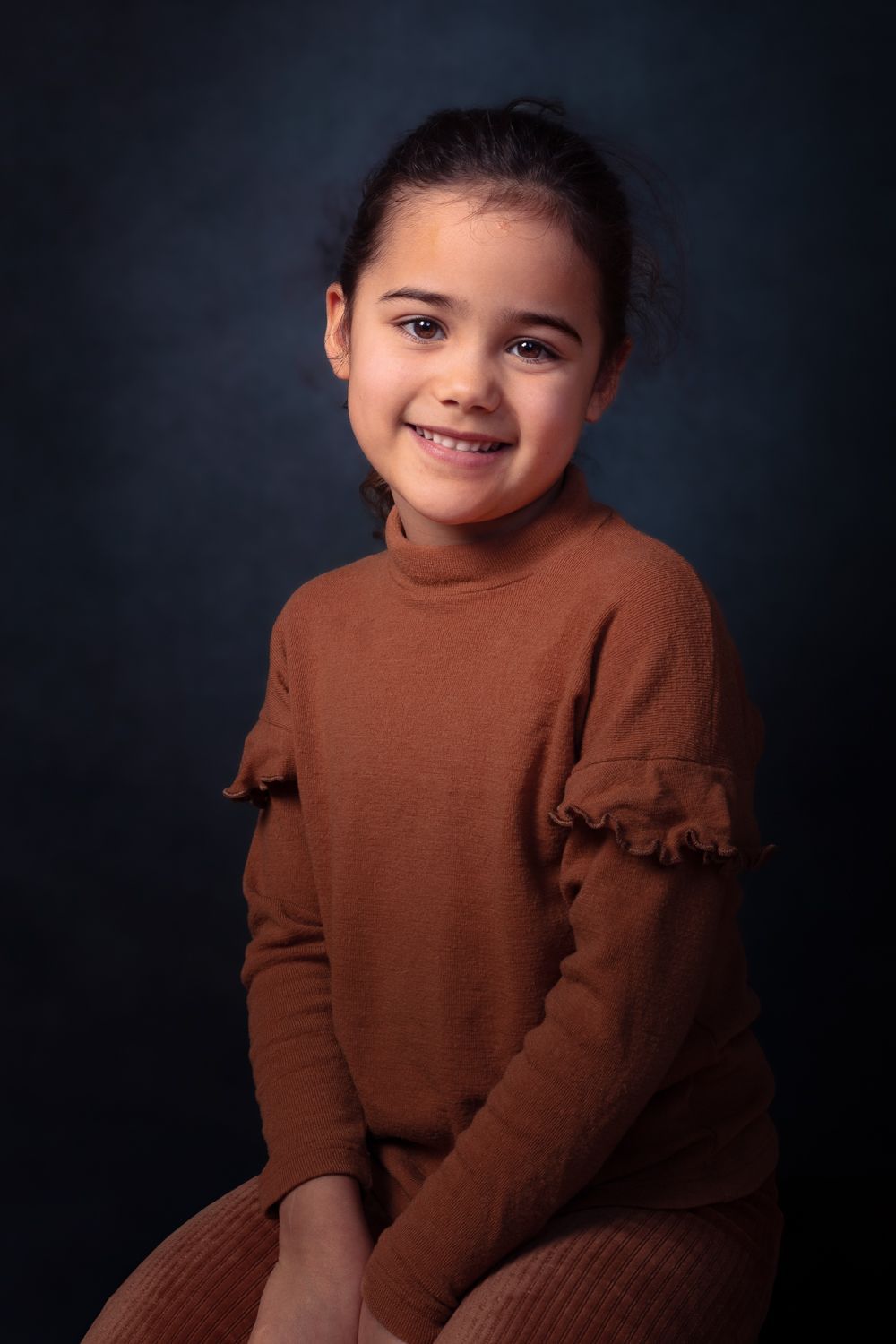 Lachend meisje, gekleed in een bruine trui, tegen een donkerblauwe achtergrond.