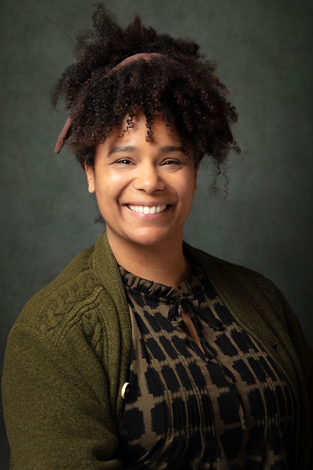 Een vrouw met krullend haar lacht, terwijl ze een olijfgroen vestje draagt, tegen een donkergroene achtergrond.