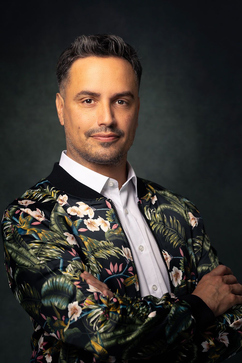 Man in bomberjack met bloemenprint, armen over elkaar, lichtjes glimlachend, tegen een donkergroene achtergrond.