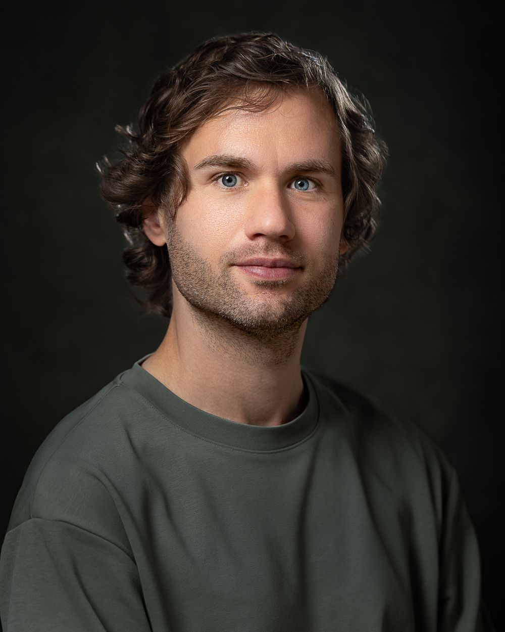 Man met een lichte huid en golvend bruin haar, gekleed in een groen shirt, kijkend in de camera. Donkere achtergrond.