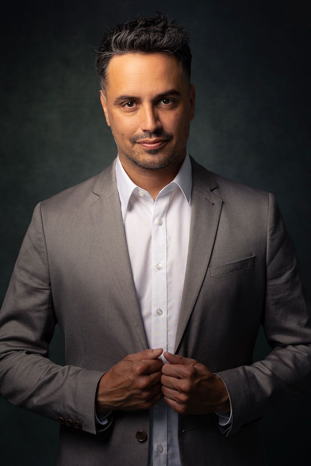 Man in grijs jasje, wit overhemd en donker haar, zijn jas vasthoudend, kijkend naar de camera. Donkergroene achtergrond.