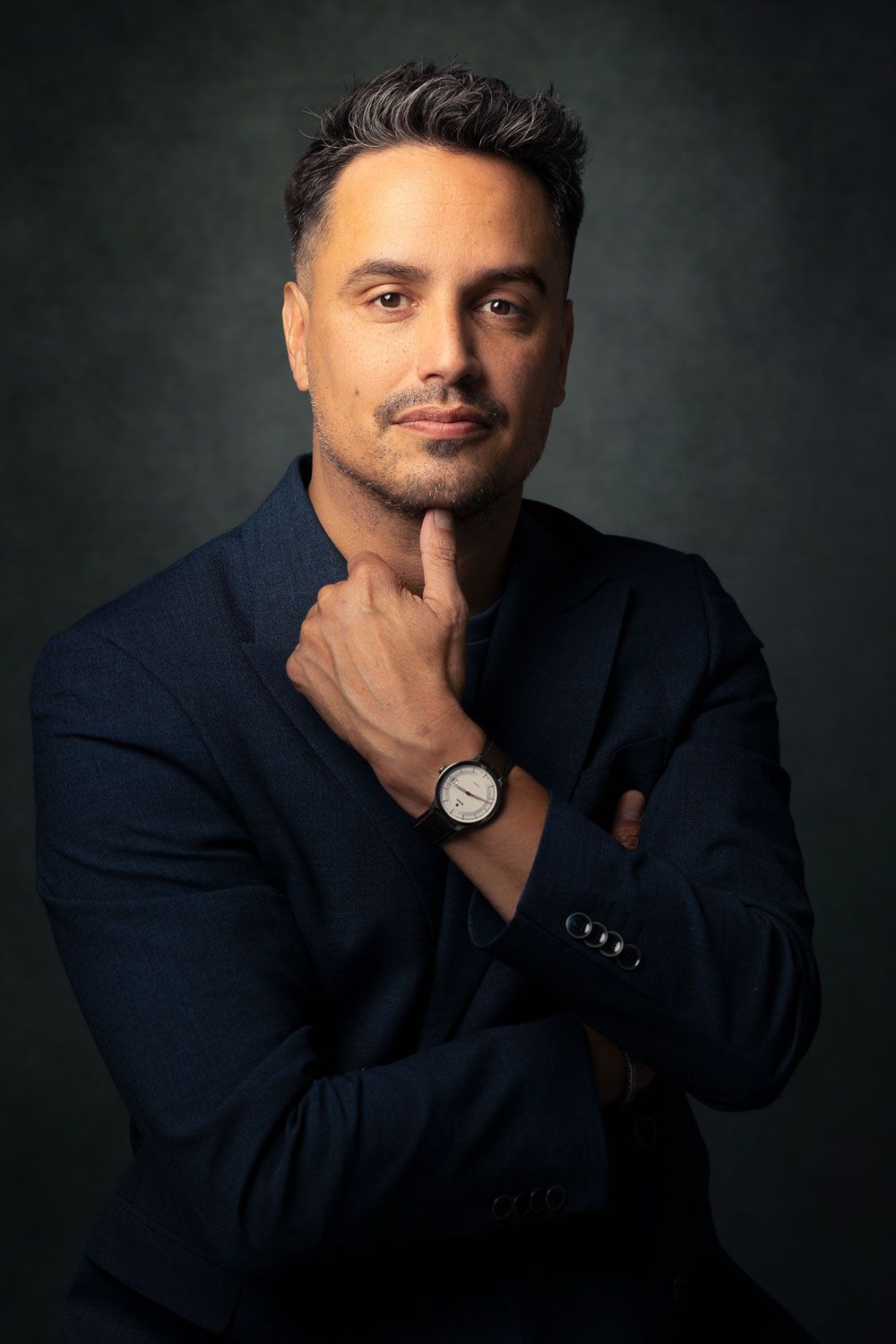 Man in een blauw pak, hand onder de kin, kijkt nadenkend in de camera, tegen een donkere achtergrond.