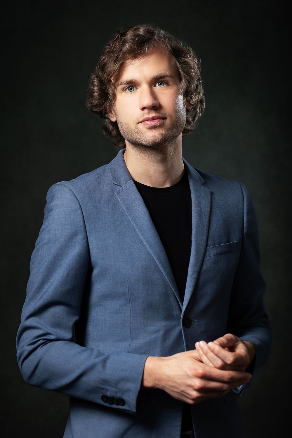 Man in blauw jasje, zwart overhemd, krullend bruin haar, handen ineengeslagen. Donkere achtergrond.