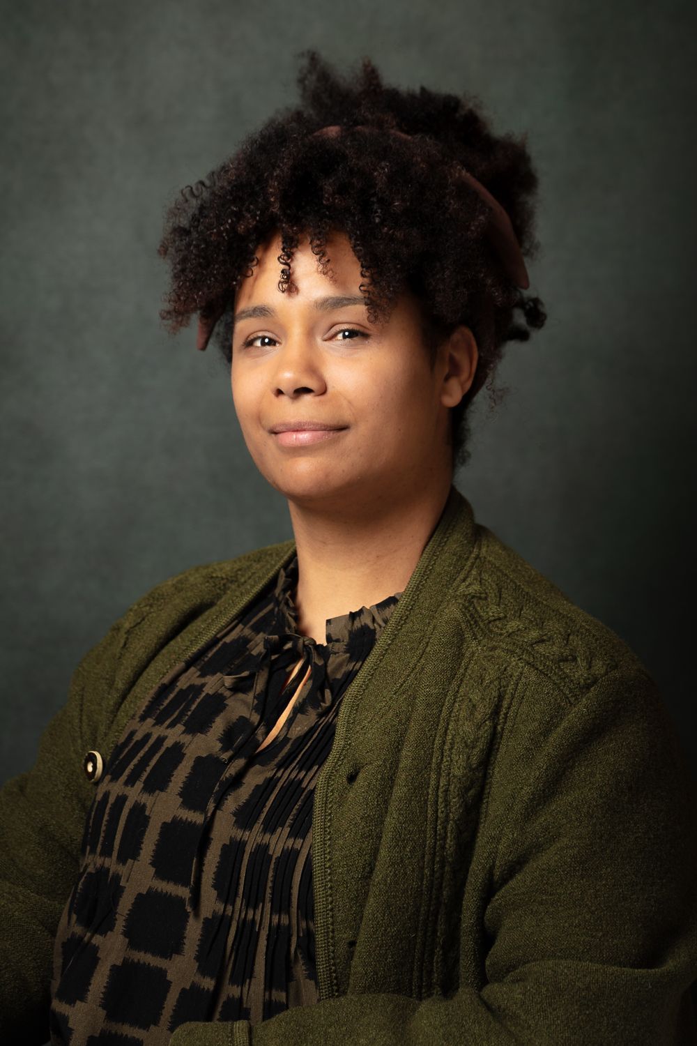 Vrouw met krullend haar in een topje met print en een groen vestje, glimlachend tegen een donkergroene achtergrond.