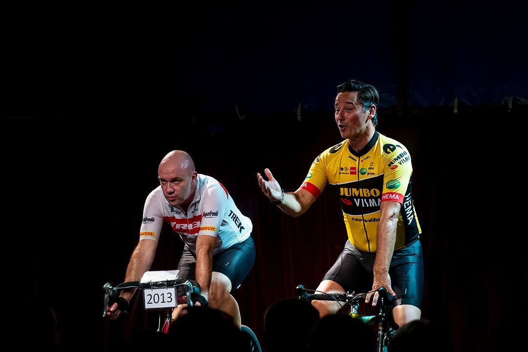 Twee fietsers op hometrainers, één maakt een gebaar, beiden zwetend; donkere omgeving.