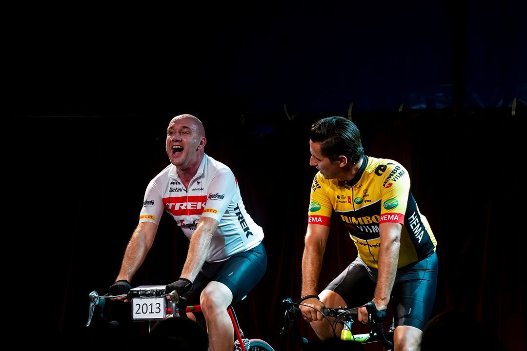 Twee fietsers op een hometrainer, één met een open mond, beiden in sportkleding, donkere achtergrond.