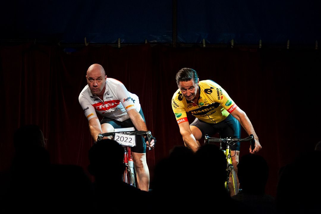 Twee wielrenners racen op hometrainers over het podium, verlicht door schijnwerpers, gezichten vol spanning.