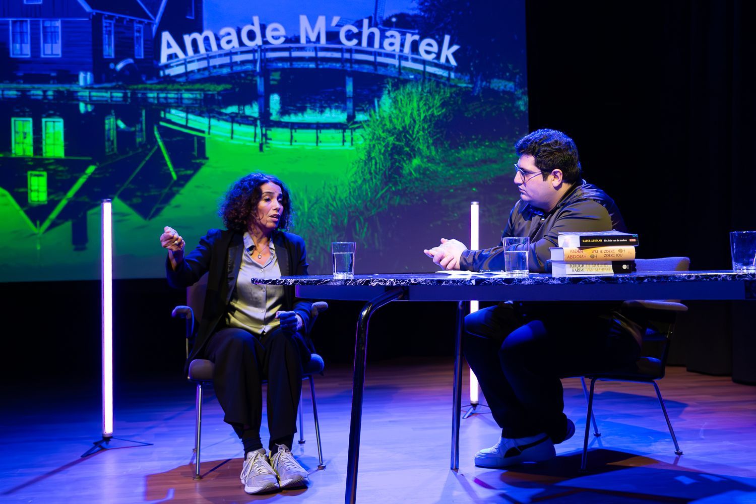 Twee mensen aan een tafel; een vrouw die een gebaar maakt, een zittende man, boeken, podium, blauw-groene achtergrond.