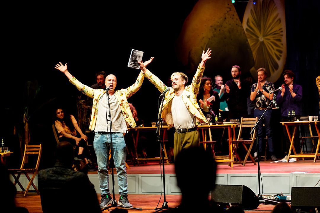 Twee mannen op het podium, met hun armen omhoog, vieren een prijs. Publiek op de achtergrond.