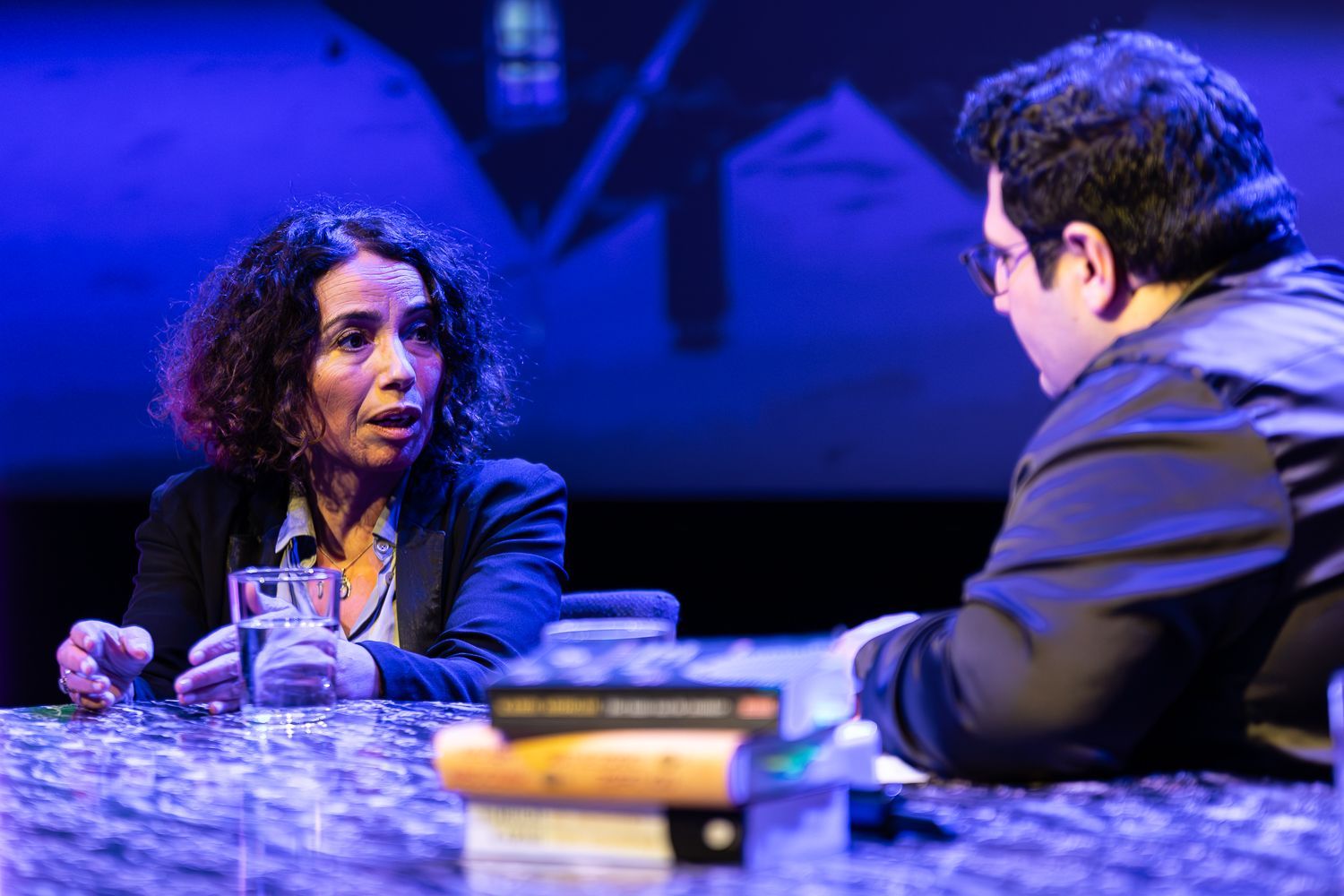 Vrouw praat met iemand buiten beeld, zittend aan een tafel met boeken. Blauwe verlichting, toneelachtige setting.