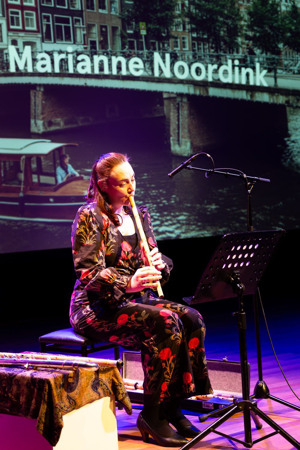 fluitist die op een houten fluit speelt, zittend op een podium, op de achtergrond het Amsterdamse kanaal.