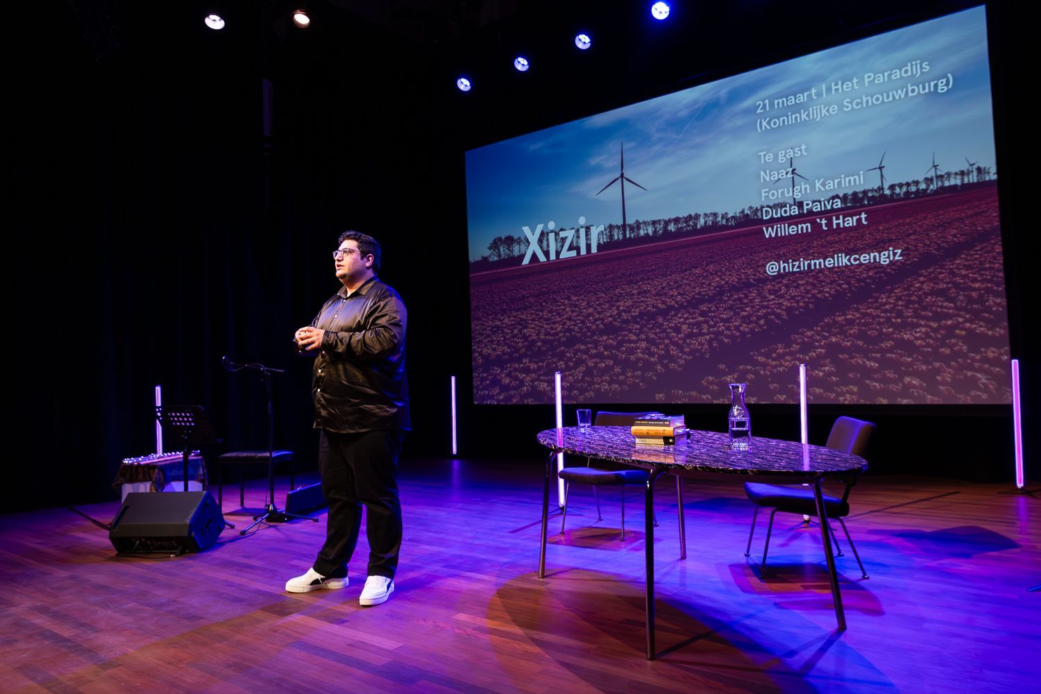 Een persoon op het podium spreekt. Achter hem verschijnt een groot scherm met daarop tekst en een afbeelding van een veld.