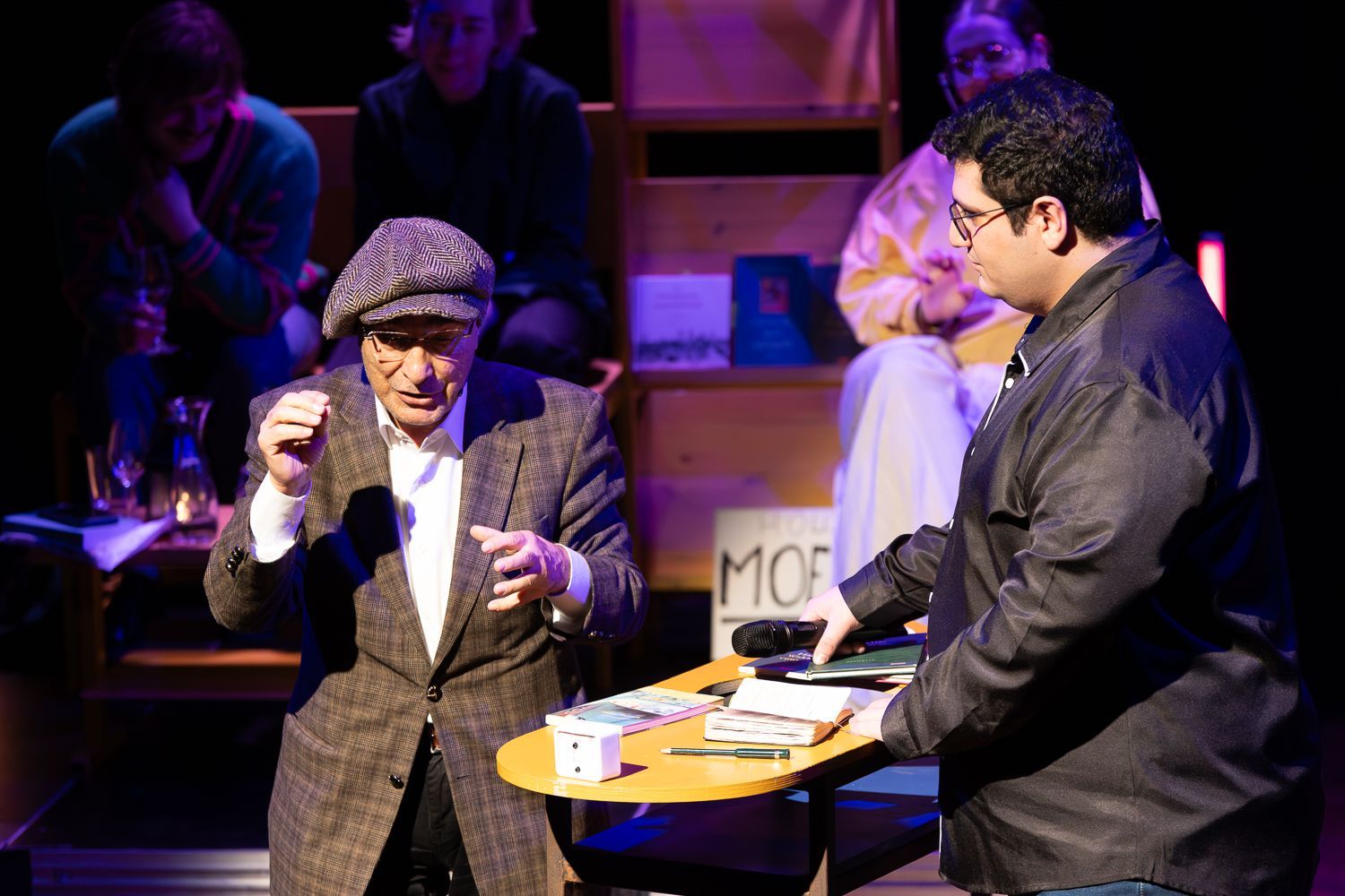 Een man in een tweedjasje wijst naar een ronde tafel waar een andere man voorwerpen vasthoudt. Het publiek zit erachter.