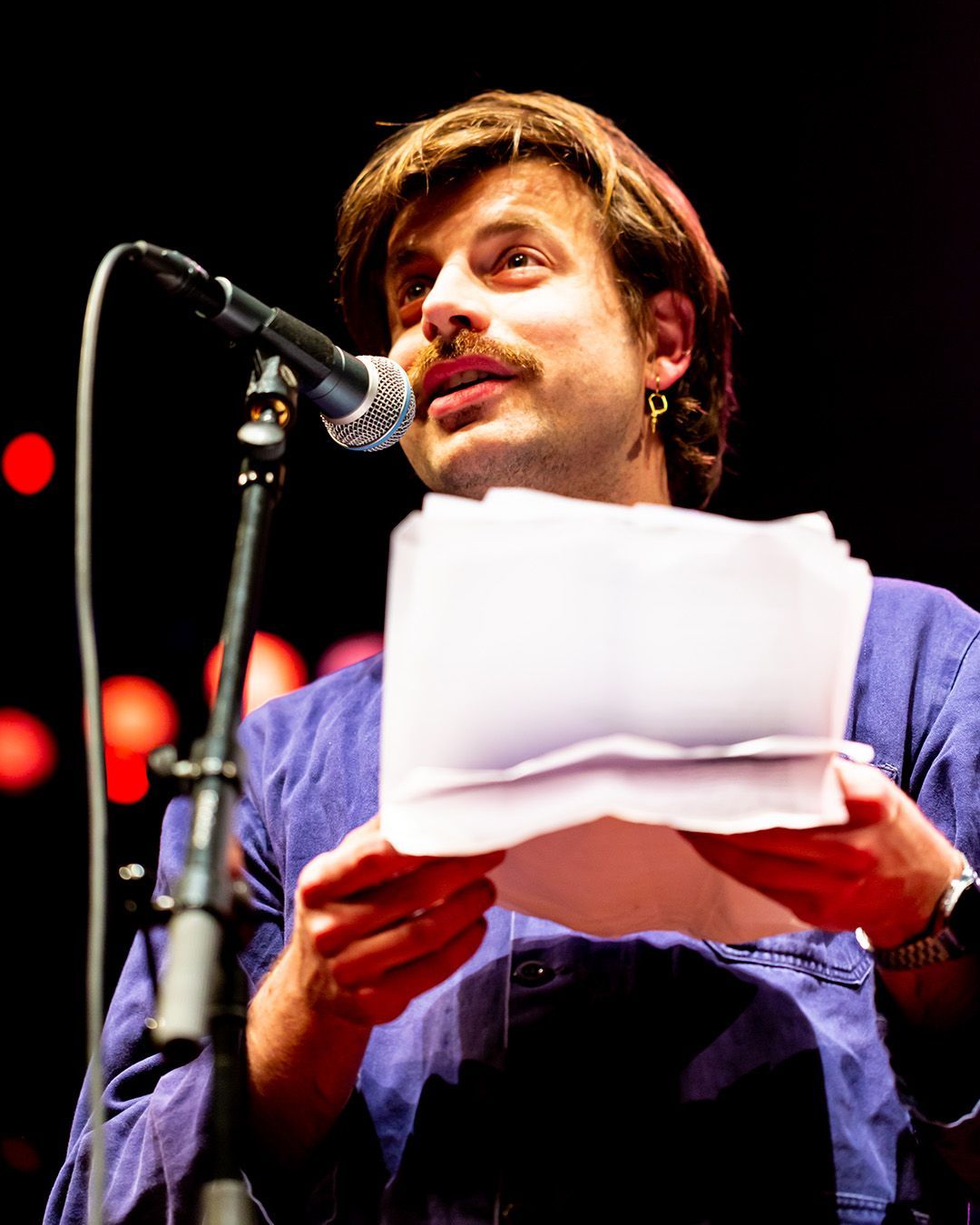 Man met snor spreekt in een microfoon, houdt papieren vast. Hij draagt een blauw shirt op een podium.