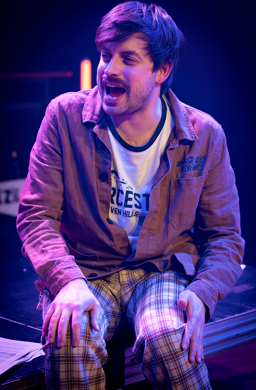 Man met bruin haar, gekleed in shirt, jas en geruite broek, kijkt verbaasd, verlicht op het podium.