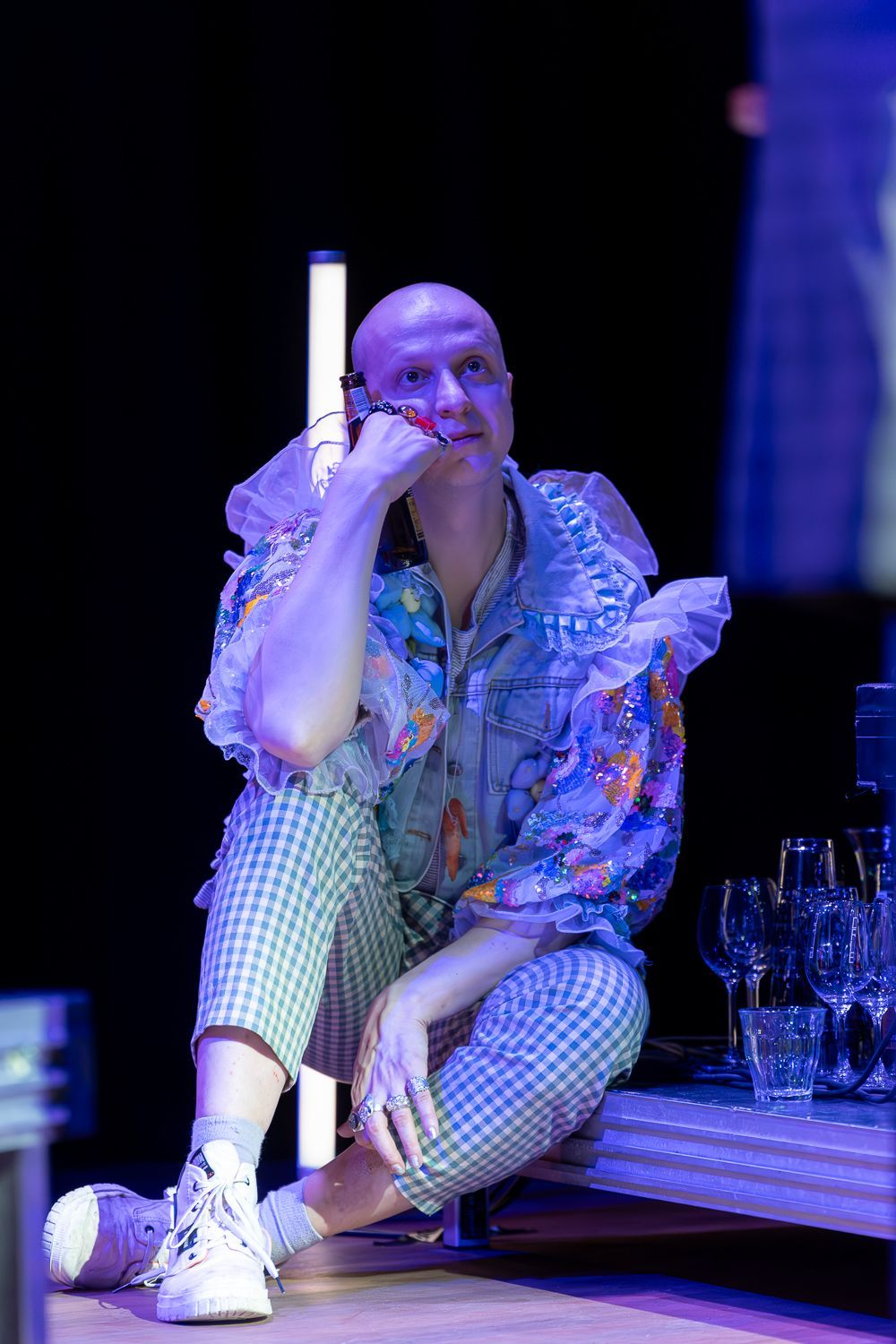 Een kale persoon draagt een kleurrijk shirt met ruches, een geruite broek en witte sneakers. Hij zit te telefoneren.