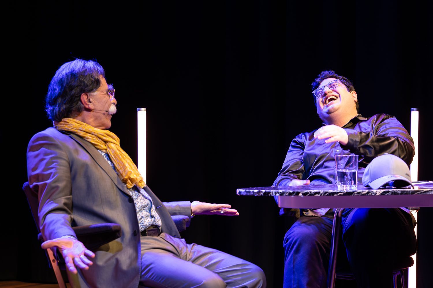 Twee mannen zitten aan een klein tafeltje, een met een snor en een sjaal, lachend. Toneeldecor, donkere achtergrond.