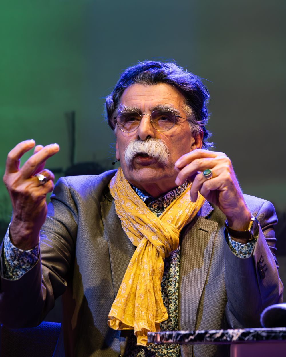 Man met snor, gekleed in een blazer, een bloemetjeshemd, een gele sjaal en ringen, aan het praten.