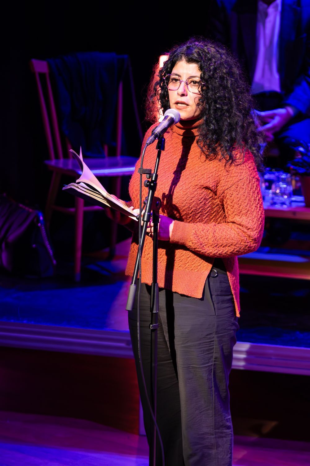 Vrouw met krullend haar spreekt in een microfoon, met tekst in haar hand. Ze draagt een oranje trui en een donkere broek.