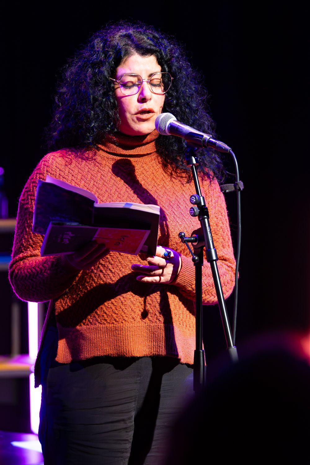 Een vrouw draagt een roestkleurige trui en leest via een microfoon op een podium voor uit een boek.