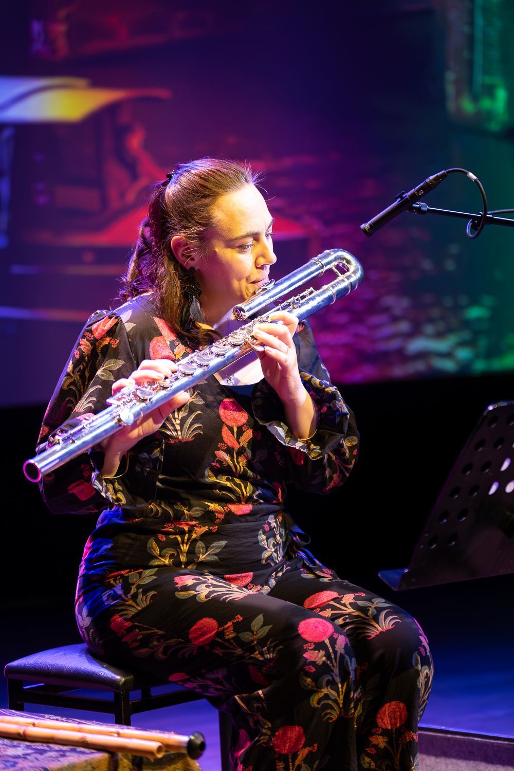 Een vrouw zit op een podium en bespeelt een zilveren fluit. Ze draagt een jurk met bloemenmotief.