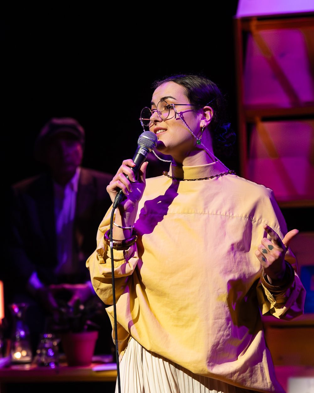 Vrouw zingt in een microfoon op het podium; geel topje, bril, donker haar, arm in de lucht.
