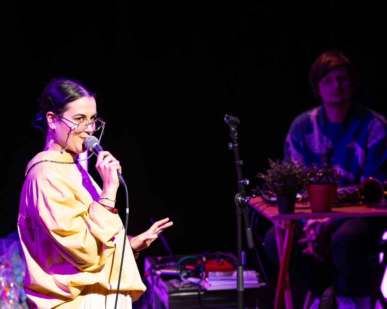 Een vrouw zingt in een microfoon op het podium; iemand met kort haar op de achtergrond. Donkere omgeving, podiumverlichting.