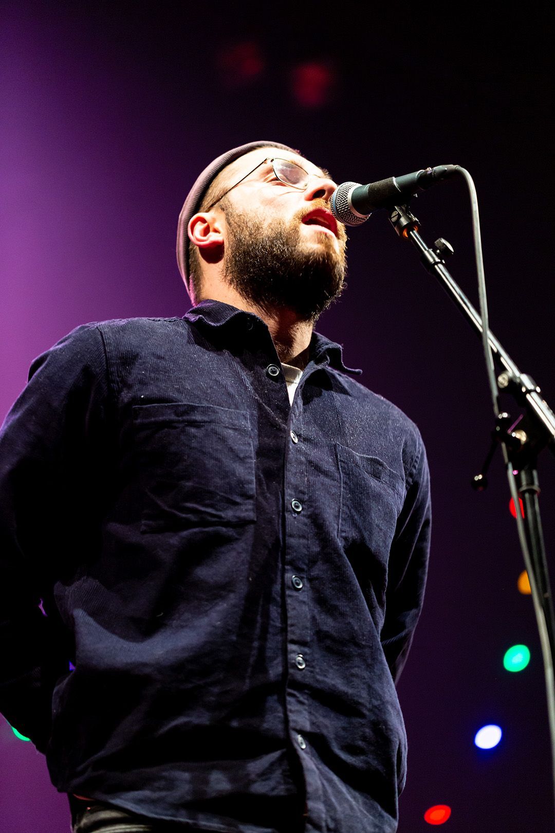 Man met bril en muts zingt in een microfoon op het podium; paarse achtergrond.