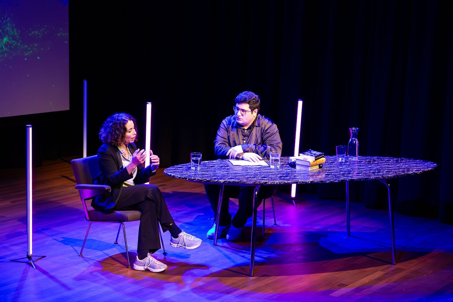 Twee mensen zitten aan een ronde tafel op een podium met neonverlichting. Eén persoon spreekt.