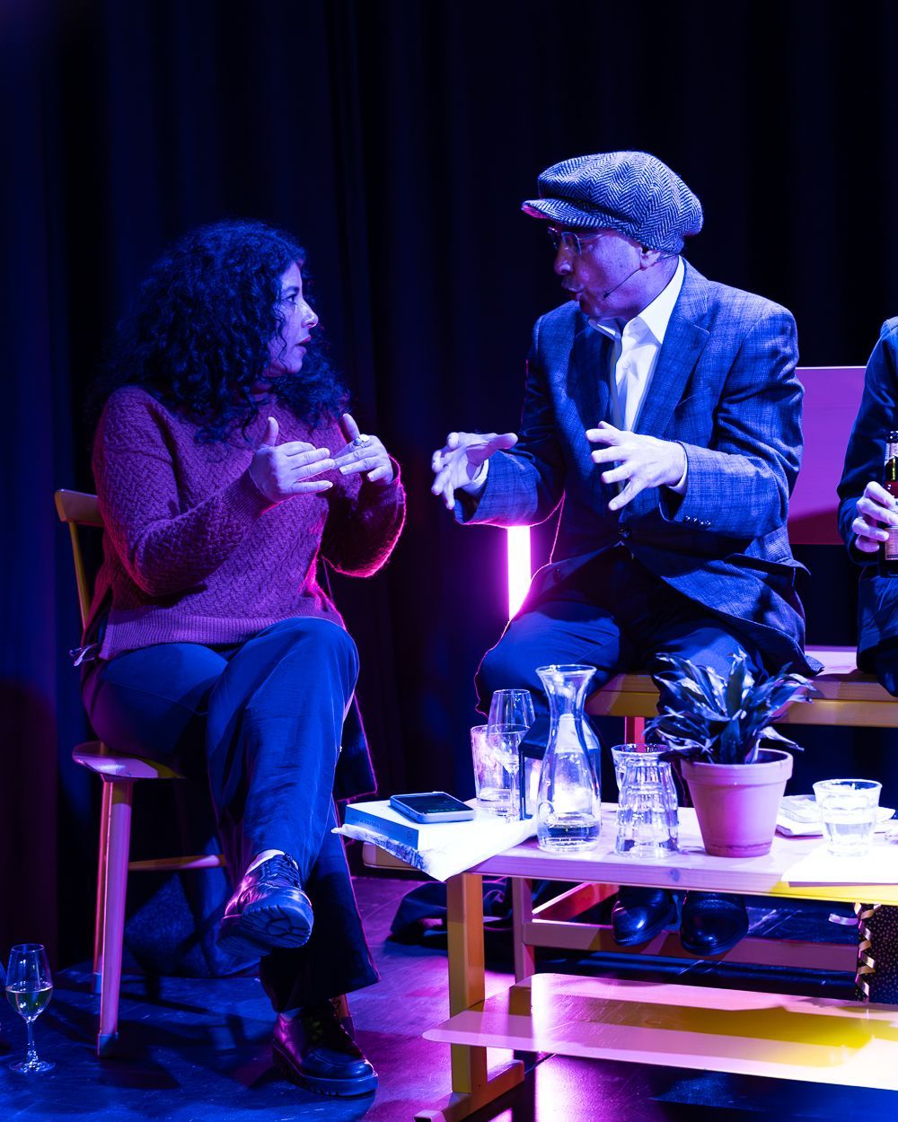 Twee mensen zitten op het podium te praten, met een tafeltje, planten en glaswerk. De verlichting is paars.