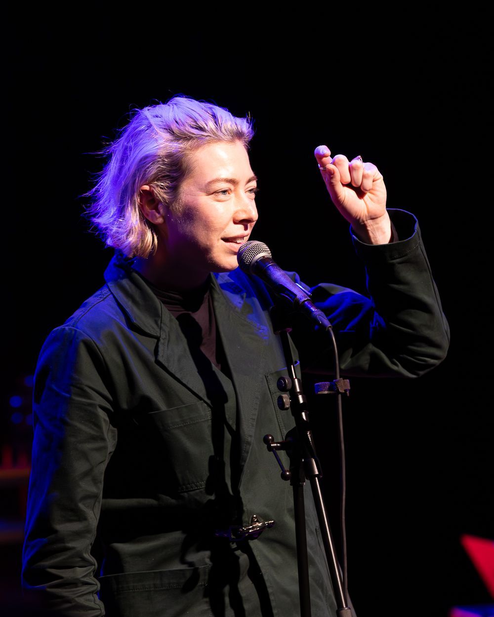 Een vrouw met blond haar houdt een microfoon vast en maakt gebaren met de rechterhand terwijl hij op een podium optreedt.