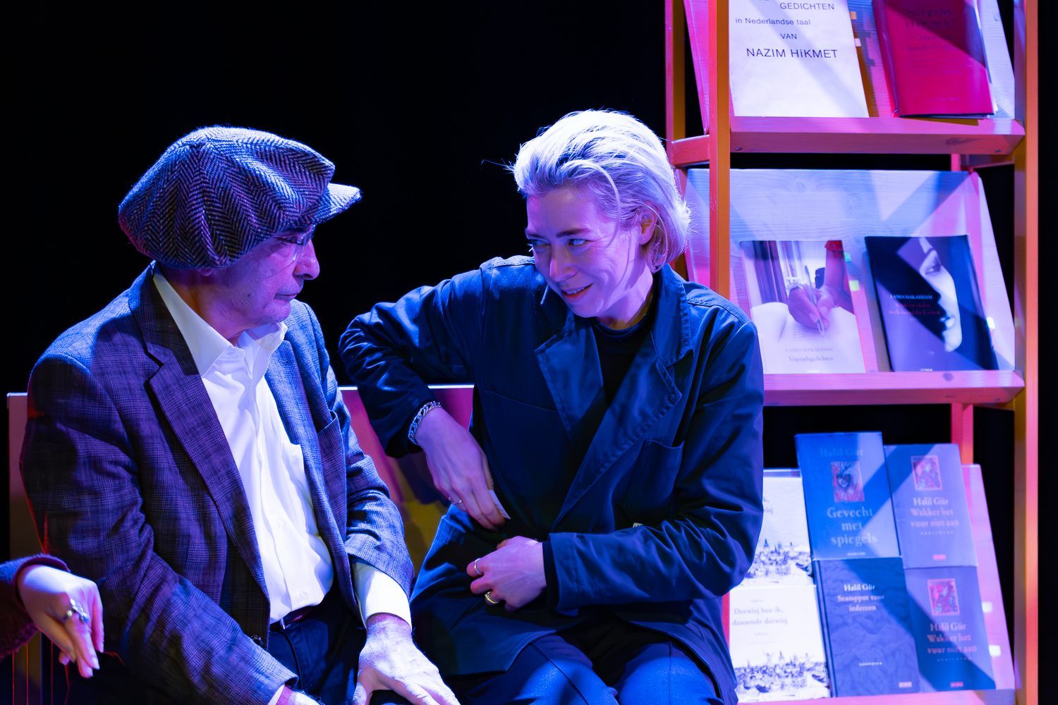 Twee mensen zitten met elkaar te praten bij een boekenplank. De een draagt een pet en een jas. De ander maakt gebaren.