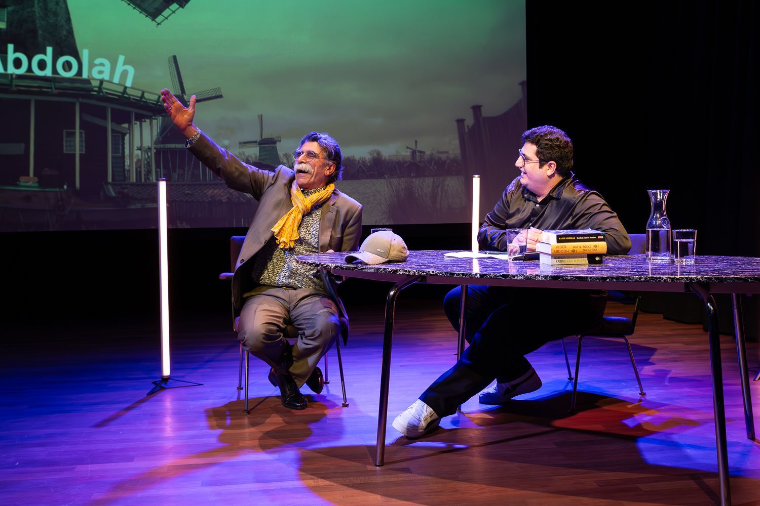 Twee mannen op het podium. De een gebaart, de ander zit. Tafel met boeken. Podiumdecor met tekst en een structuur.