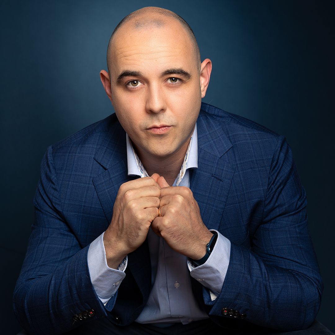 Kale man in blauw jasje en overhemd met open kraag; vuisten onder zijn kin gebald; donkerblauwe achtergrond.