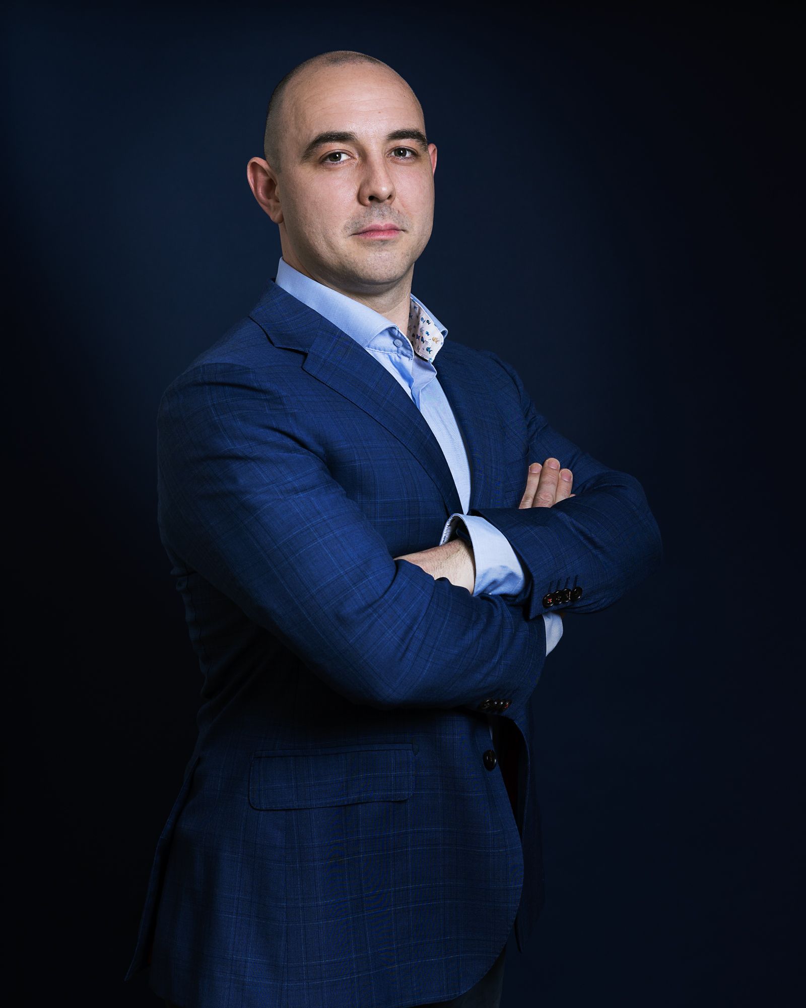 Man in een blauw jasje, met gekruiste armen, kijkt de toeschouwer aan tegen een donkere achtergrond.