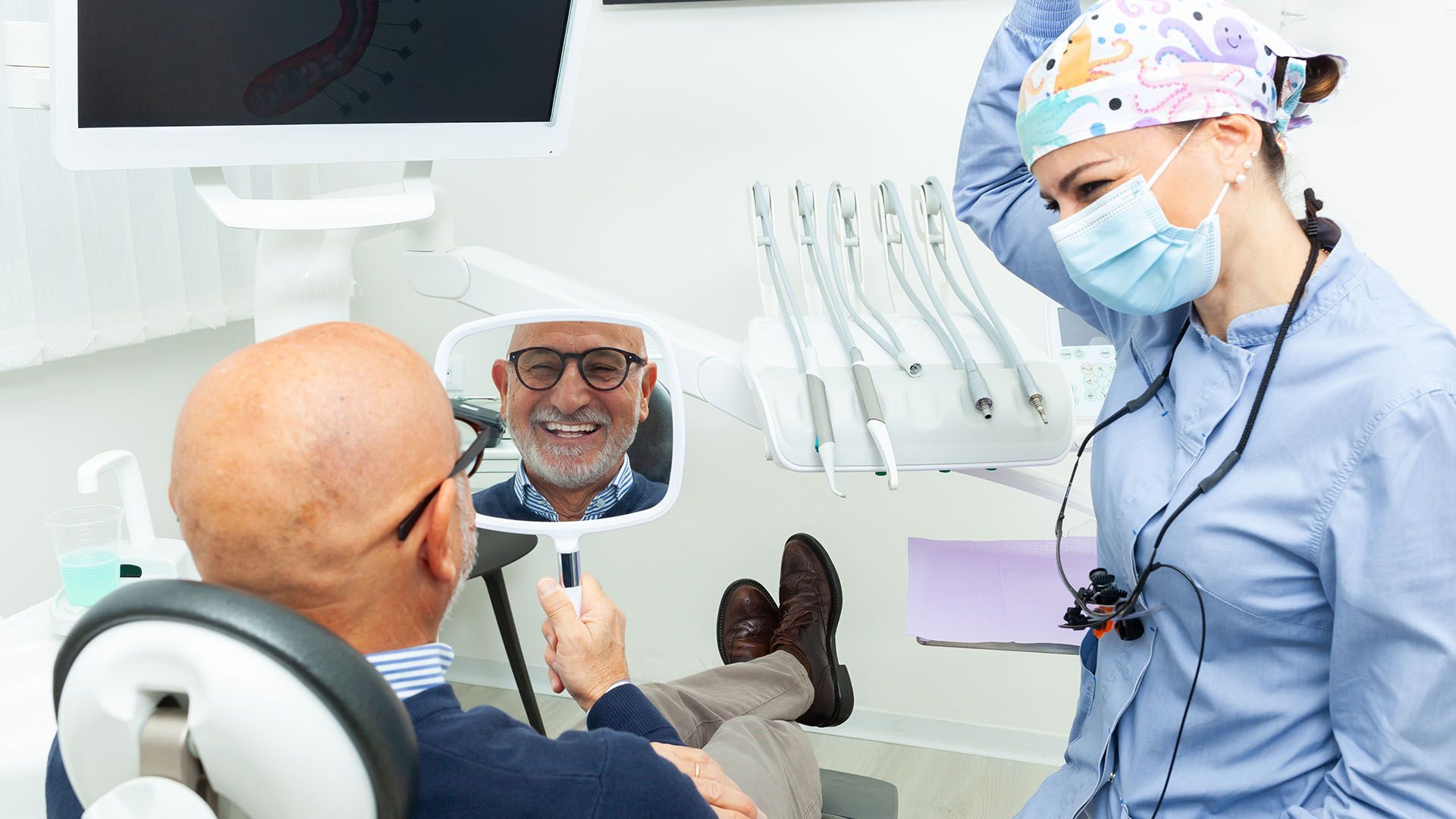 A dentist examines a smiling patient's teeth in a dental chair, using a mirror. The dentist wears a mask and scrubs, while the patient looks pleased.