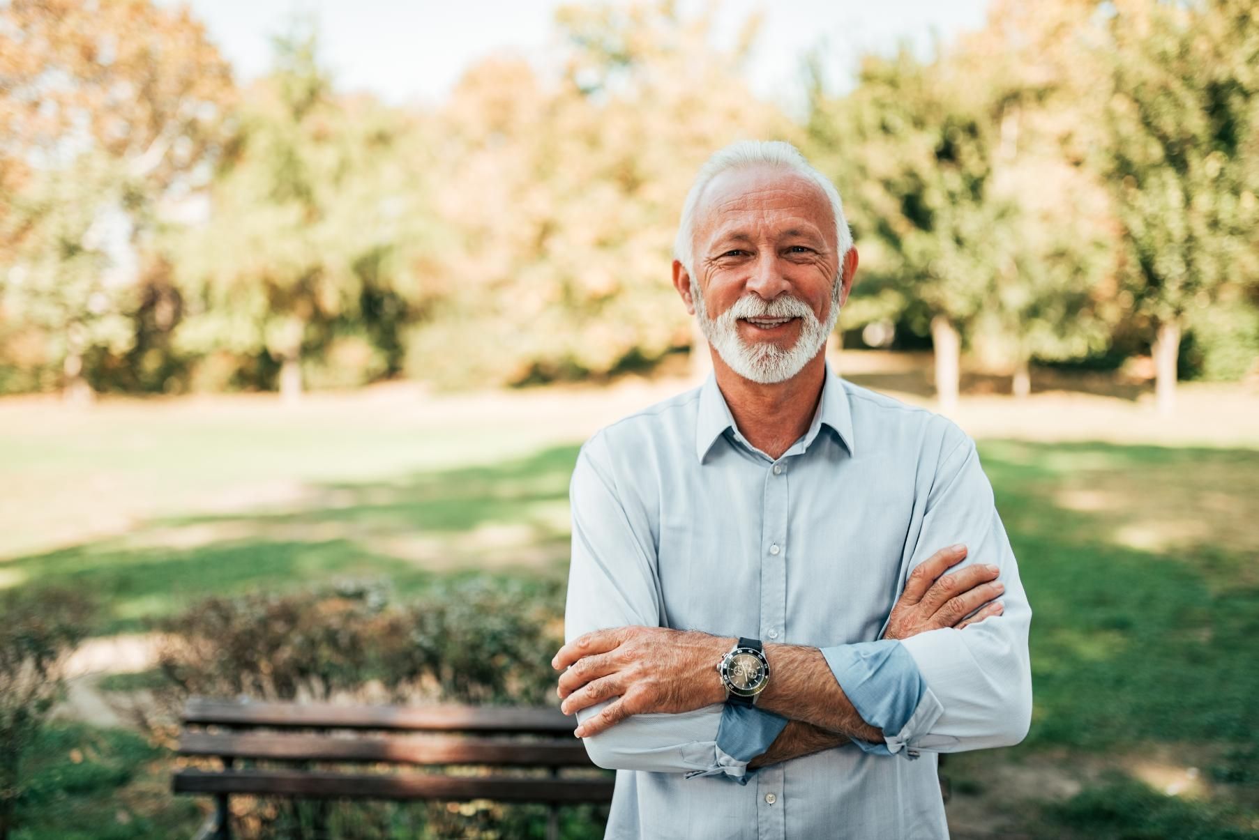 An elderly man is standing in front of a park bench with his arms crossed.