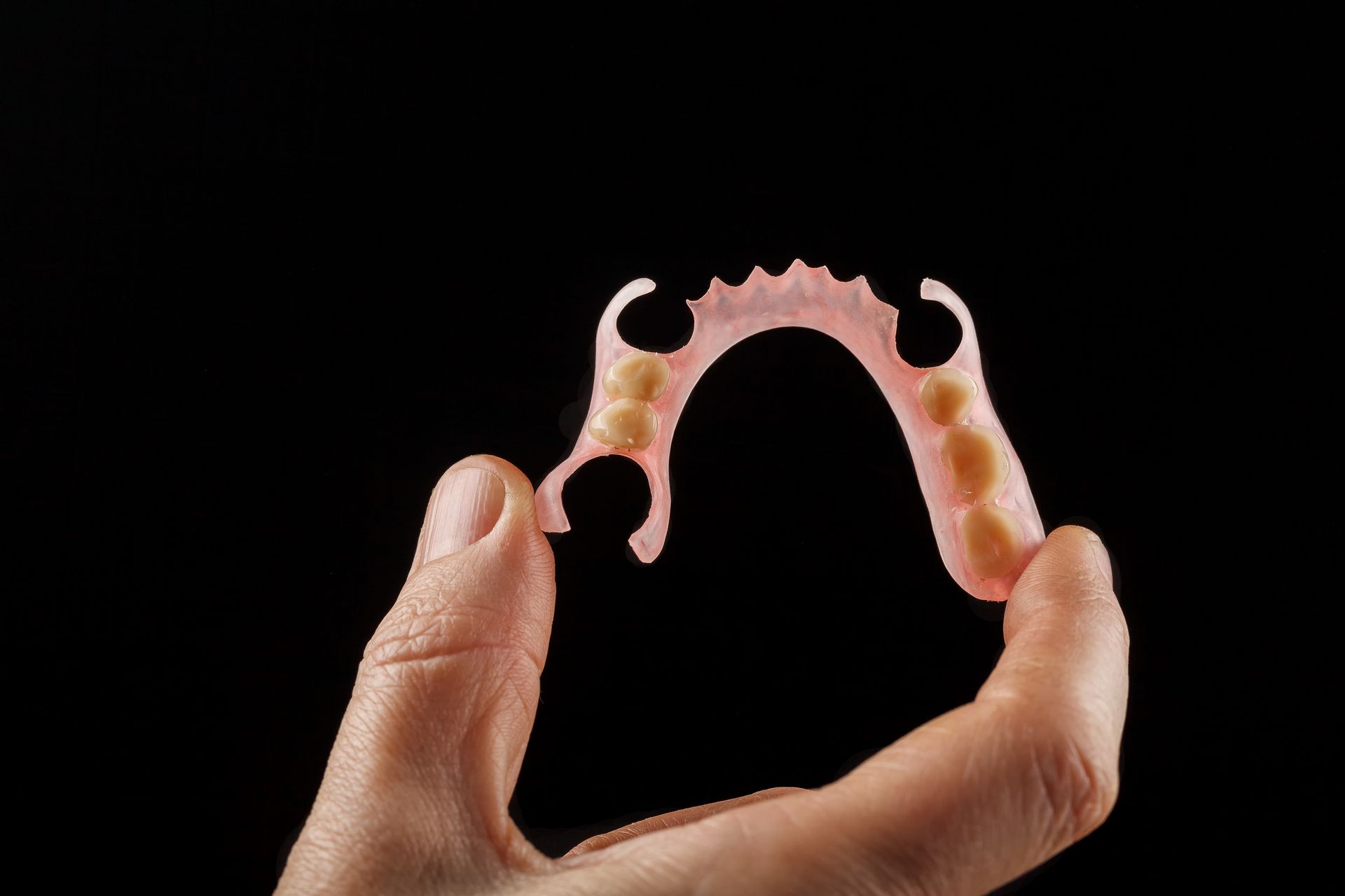 Hand holding a partial denture with pink plastic and beige artificial teeth against a black background.