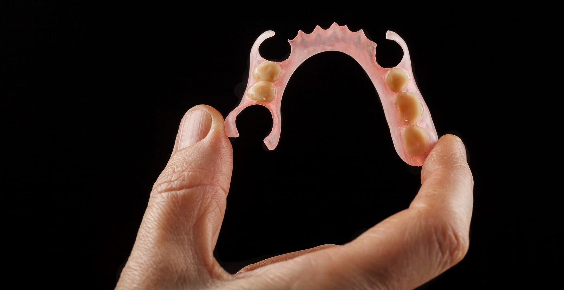 Hand holding a partial denture, pink and beige, against a black background.