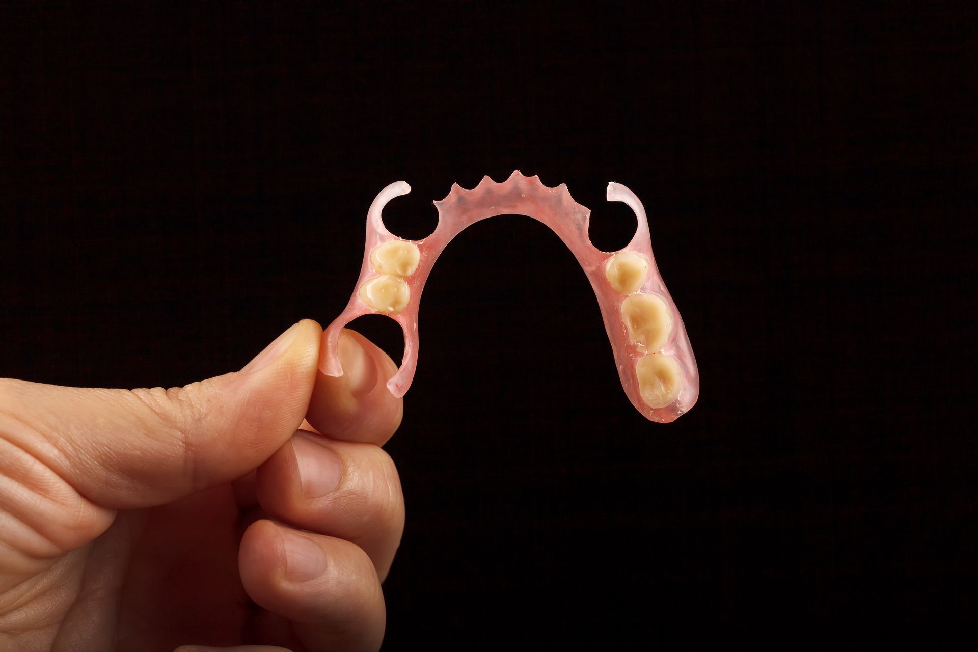 Hand holding a partial denture with pink base and tan teeth against a black background.